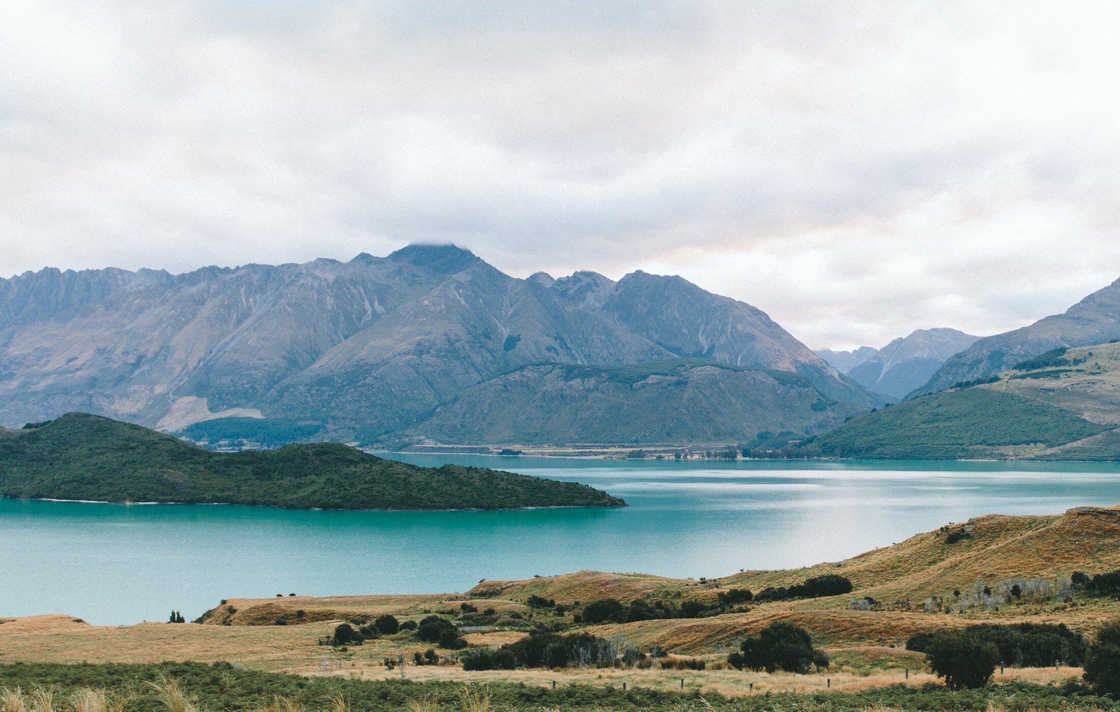 Scenic landscape with mountains, a lake, and rolling hills with sparse vegetation under cloudy sky.