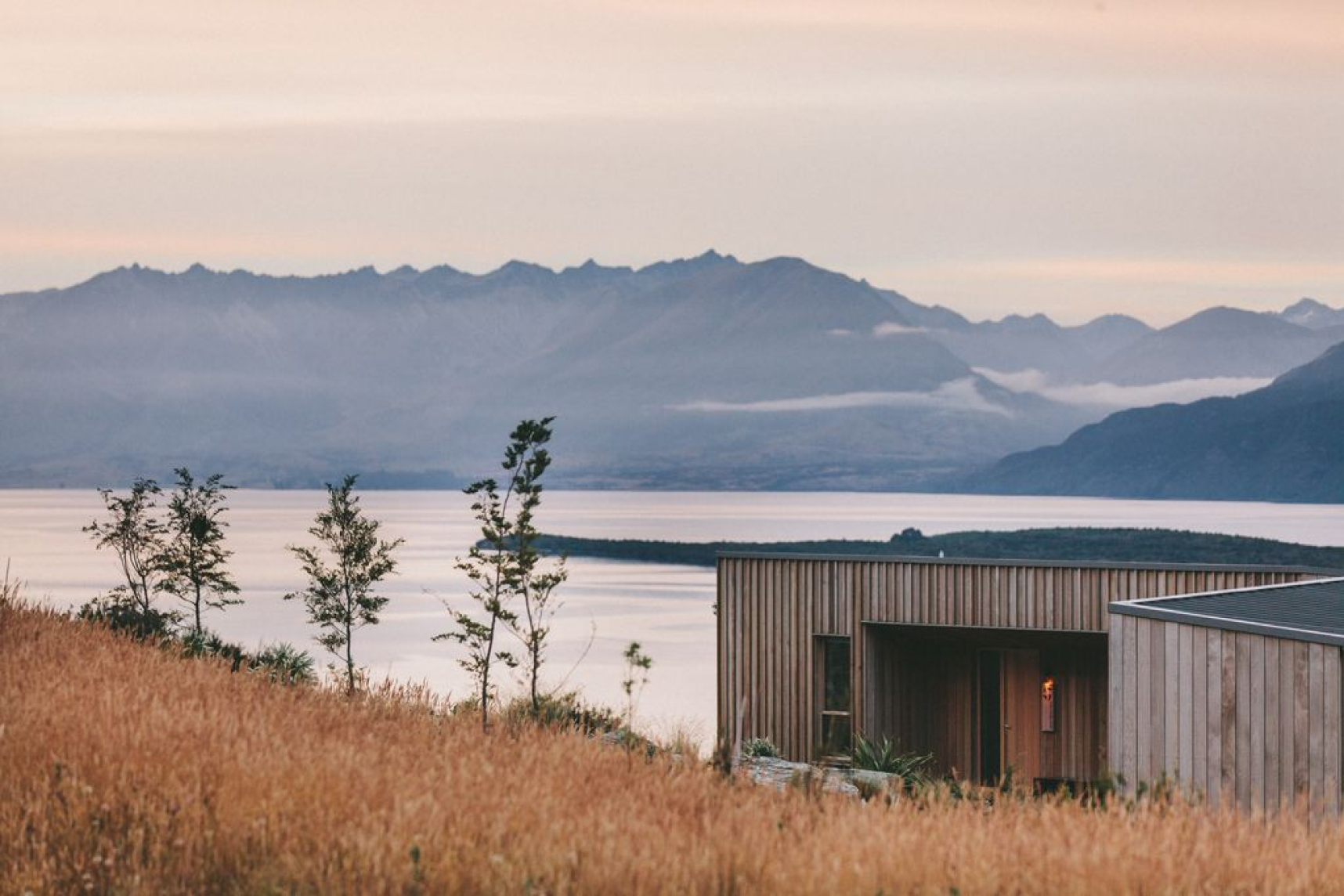 A modern wooden house with a flat roof, situated in a field of tall, golden grass, overlooking a body of water with mountains in the background under a pastel-colored sky.